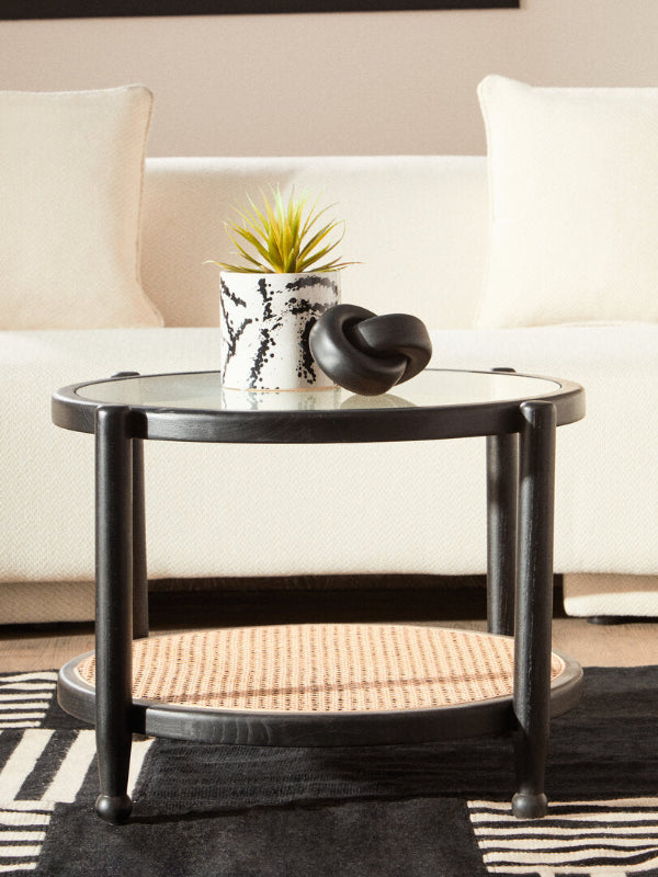 Black coffee table with a glass top and woven bottom, featuring a plant and decorative item, in a living room setting.