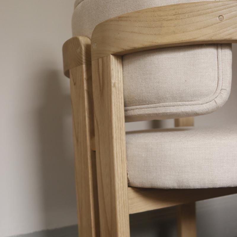 Close-up of a light natural wood chair frame with cream-colored fabric cushions, showing the armrest and seat details against a neutral wall.