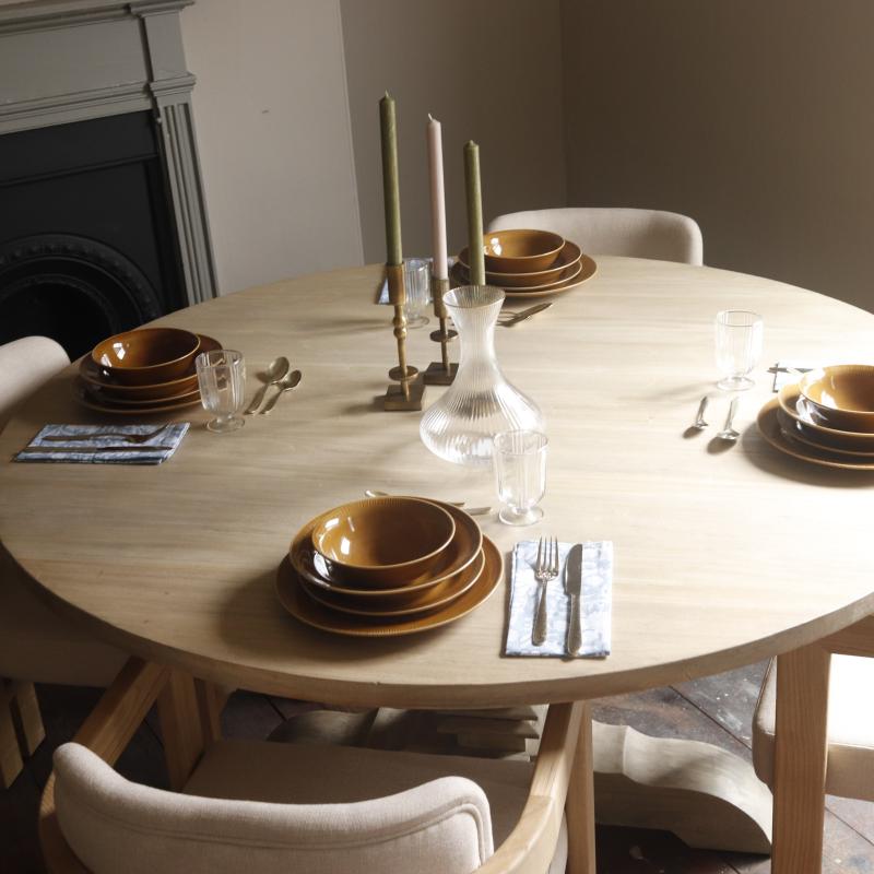 A round light wood dining table set with beige upholstered chairs, stacked amber-brown ceramic bowls, clear drinking glasses, silver cutlery, and two tall candlesticks in a softly lit indoor room.