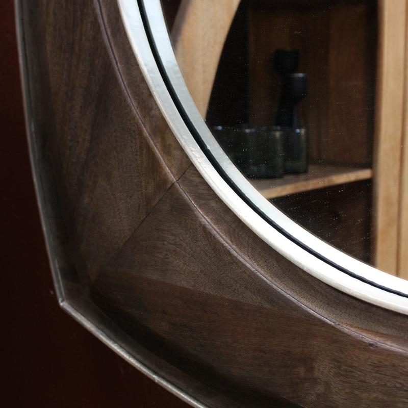 Close-up of a round wall mirror with a dark wood frame and a silver metal trim, mounted on a dark reddish-brown wall. The reflection shows wooden shelves and a candle.