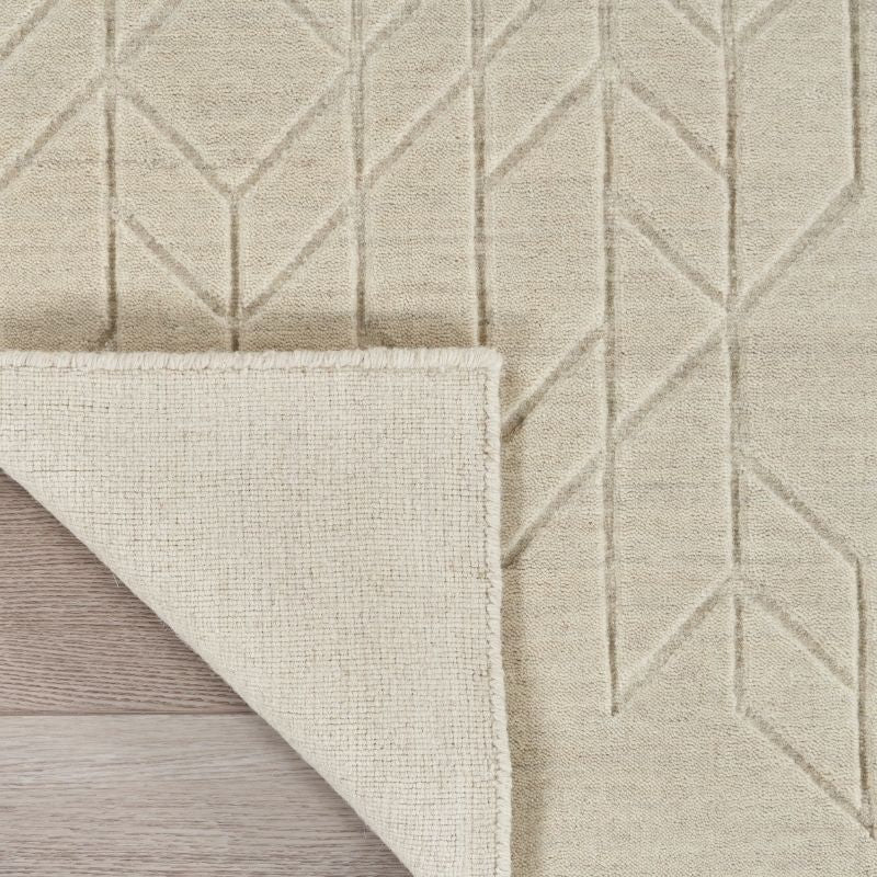 Close-up of a light beige textured rug with a geometric diamond pattern, showing the underside and folded corner over a light wood floor.