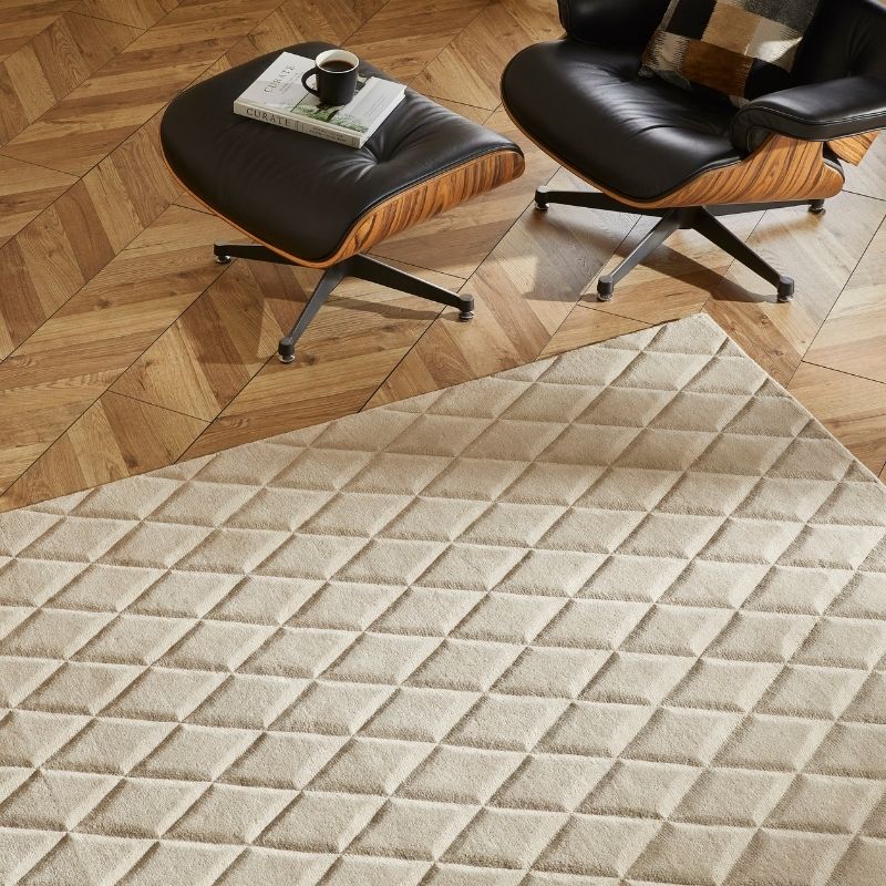 A large cream-colored area rug with a raised diamond quilted pattern, shown in a styled living room setting with two Eames-style lounge chairs and a wooden parquet floor.