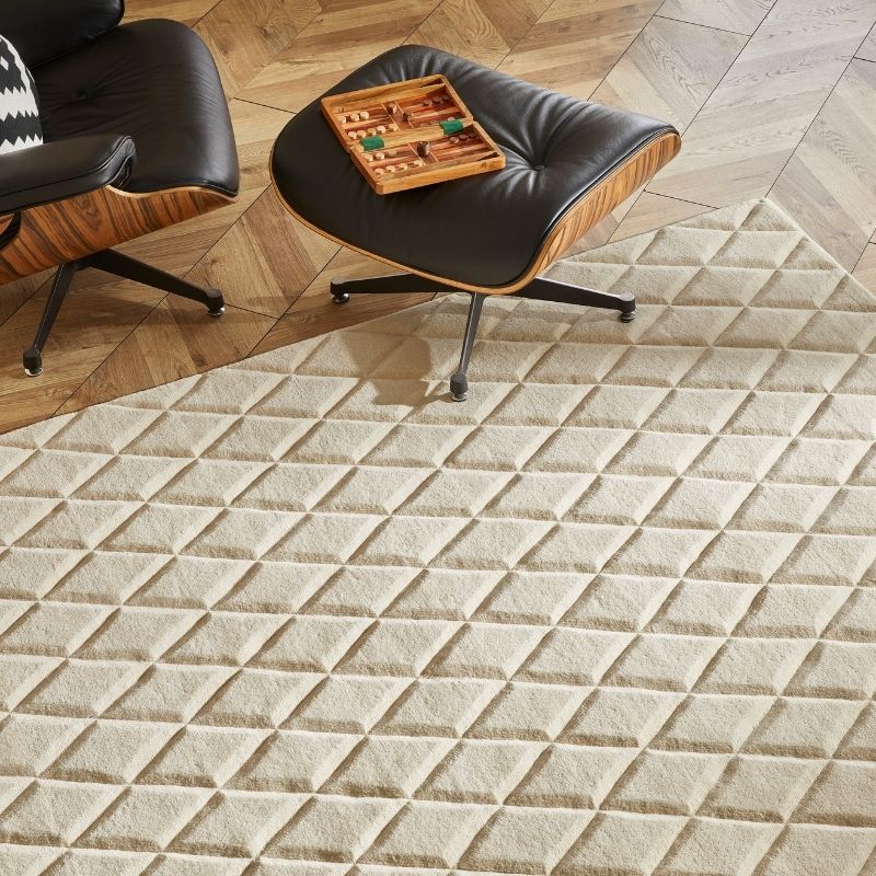A textured cream-colored area rug with a raised diamond pattern, shown in a modern living room setting alongside mid-century style chairs and a wooden floor.