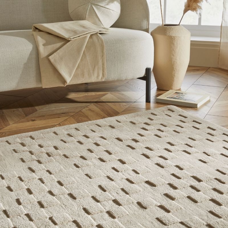 A cream-colored textured area rug with a geometric pattern of raised rectangular shapes, styled in a modern living room setting next to a beige sofa and a ceramic vase.