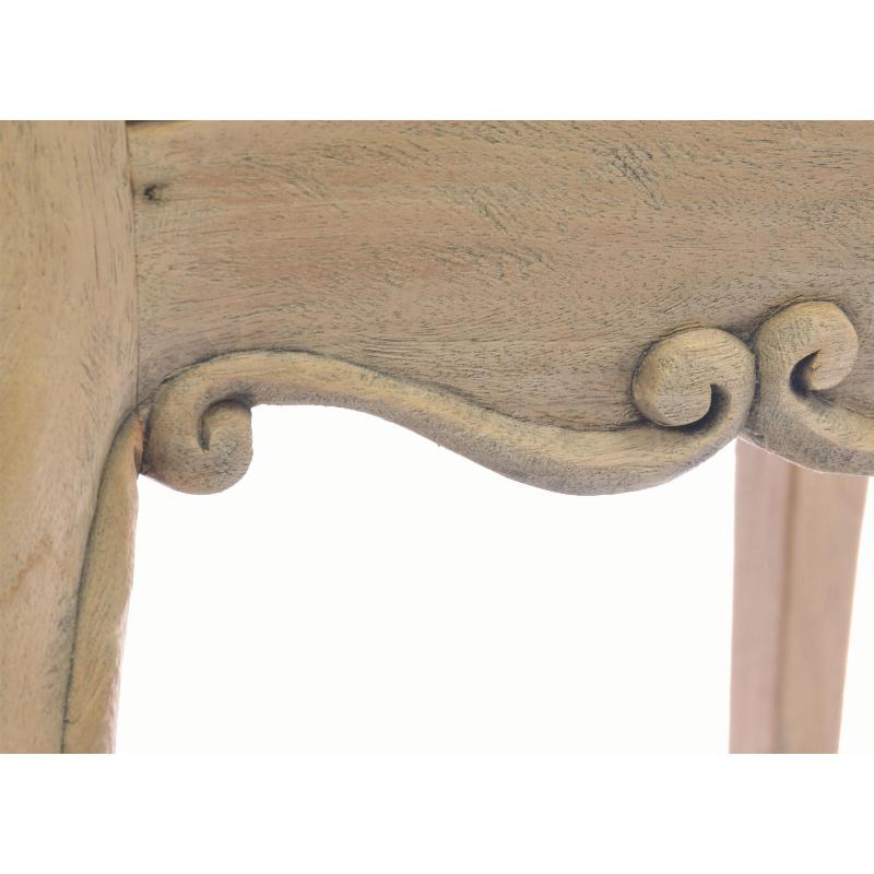 Close-up of a distressed light beige wooden table corner with ornate scrollwork carving and visible wood grain.