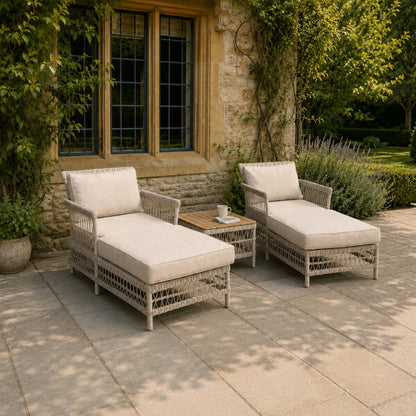 Two wicker chairs with white cushions on a stone patio in front of a stone building.