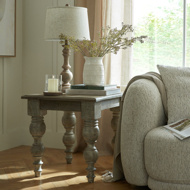 Wooden side table with lamp and vase of flowers next to a sofa in a room with natural light.