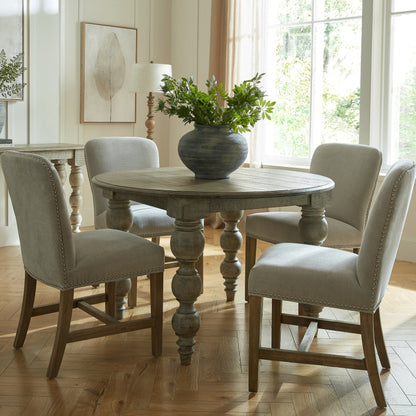 Round wooden dining table with four chairs in a bright room with large windows.