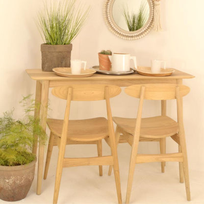 A light natural wood dining table with two matching wooden chairs, styled with white mugs, plates, and potted plants against a warm cream wall.