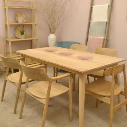 A light natural wood dining table with six matching wooden chairs, styled in a warm, minimalist room with a wooden shelf and a towel ladder.