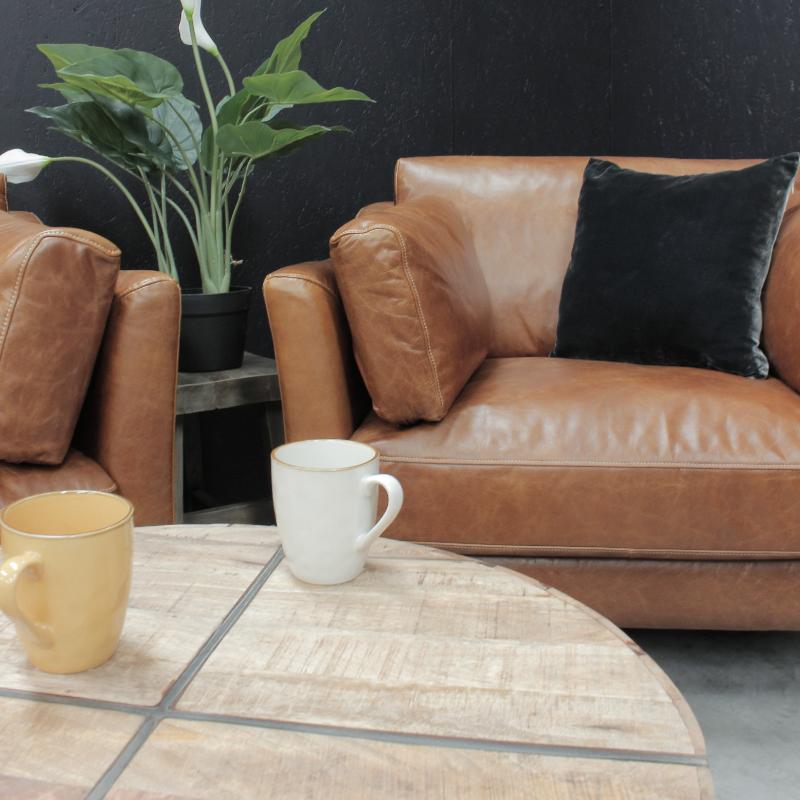 A brown leather sofa with a black throw pillow, set in a modern living room scene with a round wooden coffee table, white and yellow mugs, and a potted plant against a dark textured wall.