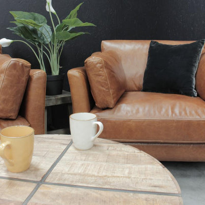A brown leather sofa with a black throw pillow, set in a modern living room scene with a round wooden coffee table, white and yellow mugs, and a potted plant against a dark textured wall.