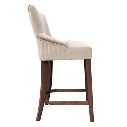 A single upholstered bar stool shown from the side against a plain white background. The seat and back are upholstered in a warm light beige fabric with vertical channel tufting and nailhead trim, supported by a dark brown wooden frame with a footrest.