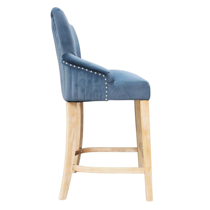 A blue velvet upholstered bar stool with a curved backrest, nailhead trim on the armrests, and light natural wood legs and footrest, shown against a plain white background.