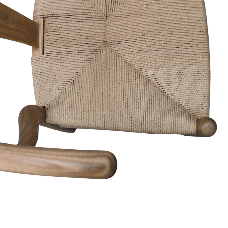 Close-up of a wooden chair seat with woven natural fiber rope, showing the wood frame and textured seat against a gray concrete floor.