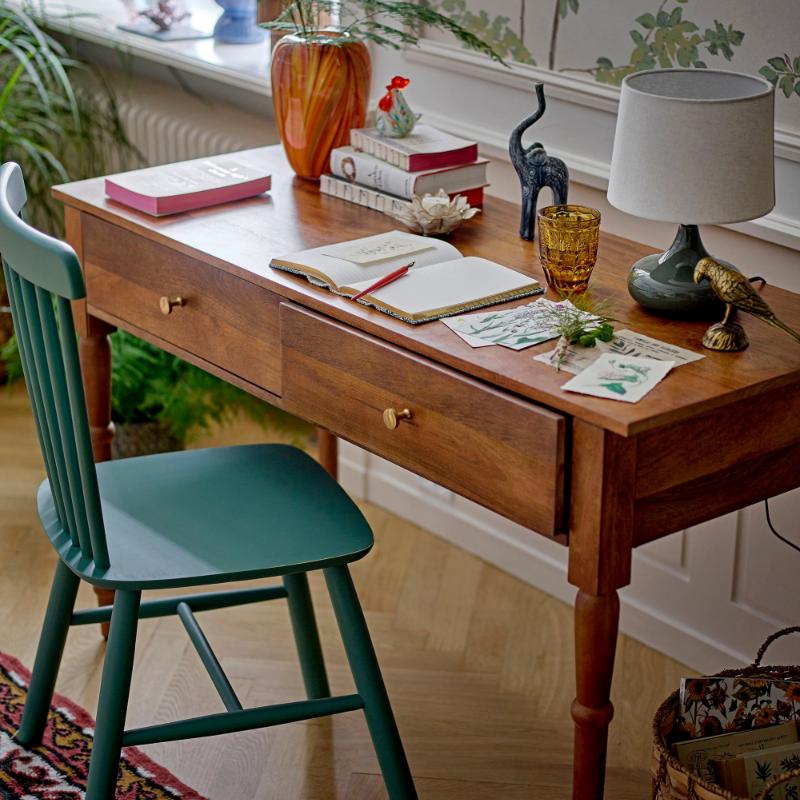 Wooden desk with various items including a lamp, books, and a green chair in a room with plants and a window.