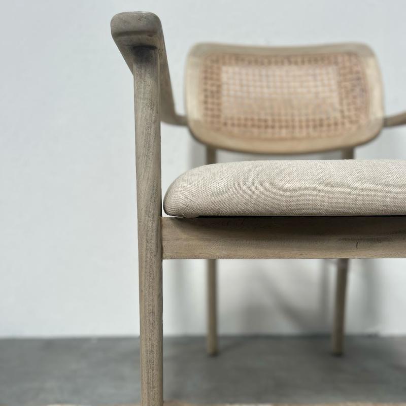 A close-up of a wooden armchair with a light beige linen seat cushion and a woven cane backrest, set against a neutral gray wall and concrete floor.