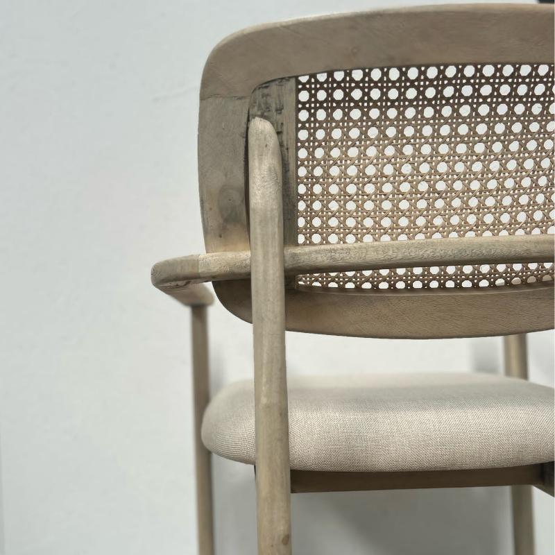 A close-up of a light beige wooden dining chair with a woven cane backrest and a textured fabric seat cushion, set against a plain light gray background.