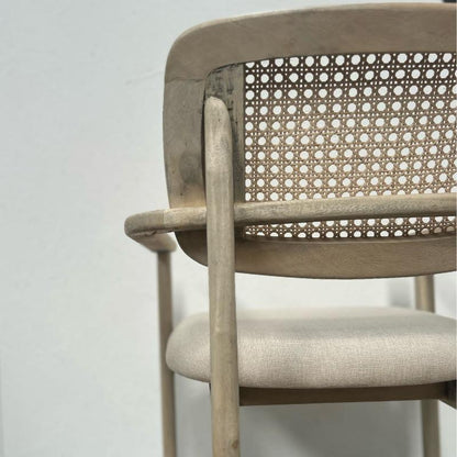 A close-up of a light beige wooden dining chair with a woven cane backrest and a textured fabric seat cushion, set against a plain light gray background.