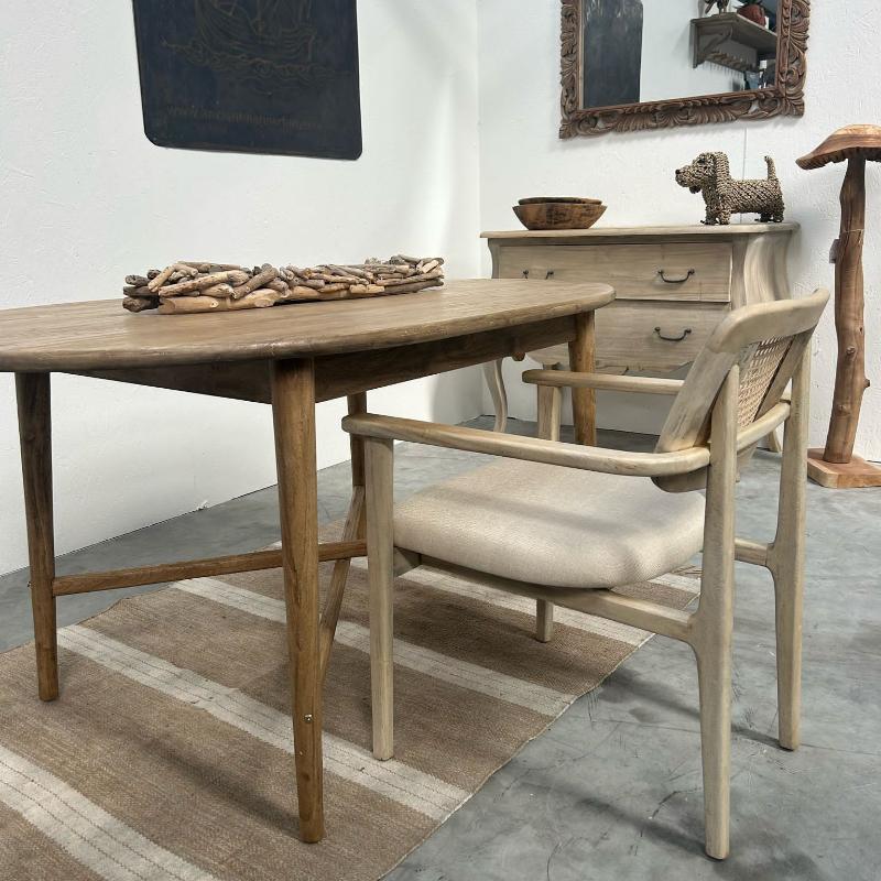 A rustic dining table with a light wood top and weathered brown legs, paired with a matching light wood armchair featuring a woven cane backrest and a beige upholstered seat. The furniture is styled on a striped beige and brown rug in a dining room setting with a wooden dresser and mirror in the background.