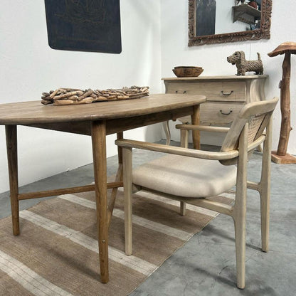 A rustic dining table with a light wood top and weathered brown legs, paired with a matching light wood armchair featuring a woven cane backrest and a beige upholstered seat. The furniture is styled on a striped beige and brown rug in a dining room setting with a wooden dresser and mirror in the background.