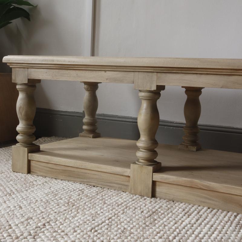 A light natural wood TV stand or low coffee table with a lower shelf, featuring turned legs and block feet, set on a textured cream rug against a neutral wall.