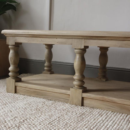 A light natural wood TV stand or low coffee table with a lower shelf, featuring turned legs and block feet, set on a textured cream rug against a neutral wall.