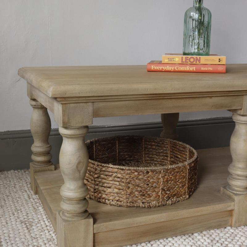 A light beige wood coffee table with turned legs and a lower shelf, styled with a woven basket, books, and a glass vase against a neutral wall.