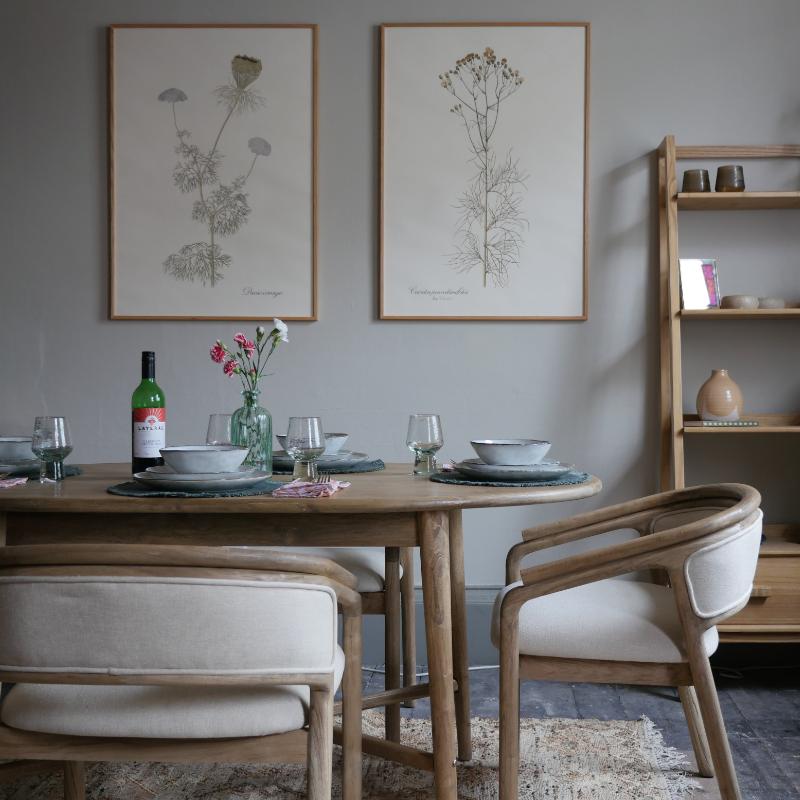A styled dining room scene featuring a round wooden table set with white plates, a green wine bottle, and a glass vase with red and white flowers. Two modern wooden armchairs with cream cushions are in the foreground, and two framed botanical prints hang on a gray wall behind the table.