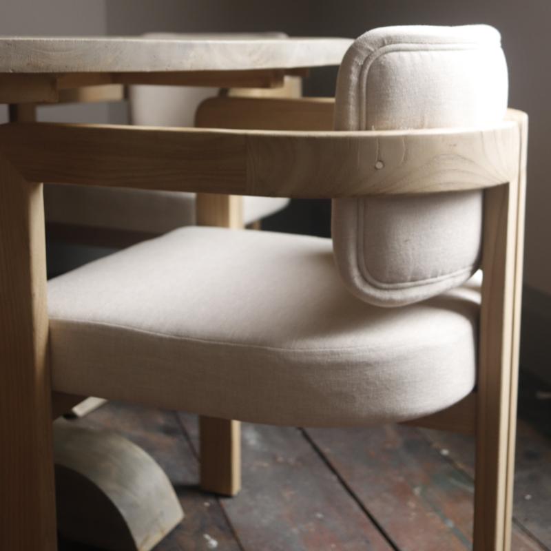A close-up of a wooden dining chair with light beige upholstered seat and back cushions, set in a rustic indoor environment with a wooden floor and table visible.