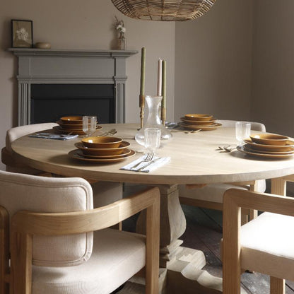 A round light wood dining table set with matching brown ceramic plates, clear drinking glasses, silver cutlery, and a glass carafe, surrounded by light wood and cream upholstered chairs in a softly lit dining room.