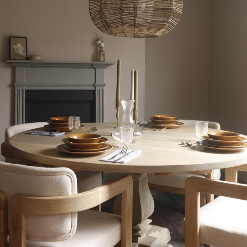 A round light wood dining table set with matching brown plates, clear drinking glasses, folded silverware, and tall candlesticks, surrounded by upholstered wooden chairs in a softly lit dining room with a fireplace and woven pendant light overhead.