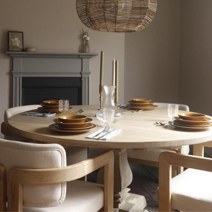 A round light wood dining table set with matching brown plates, clear drinking glasses, folded silverware, and tall candlesticks, surrounded by upholstered wooden chairs in a softly lit dining room with a fireplace and woven pendant light overhead.