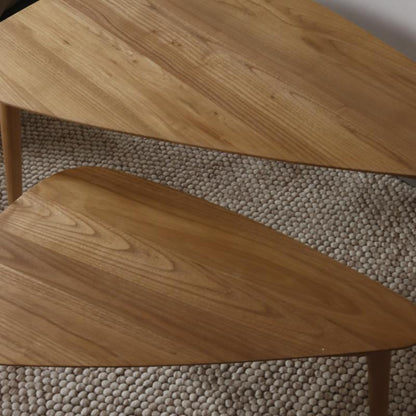 A close-up of a wooden table with a triangular top and visible wood grain, set against a textured beige pebble rug.