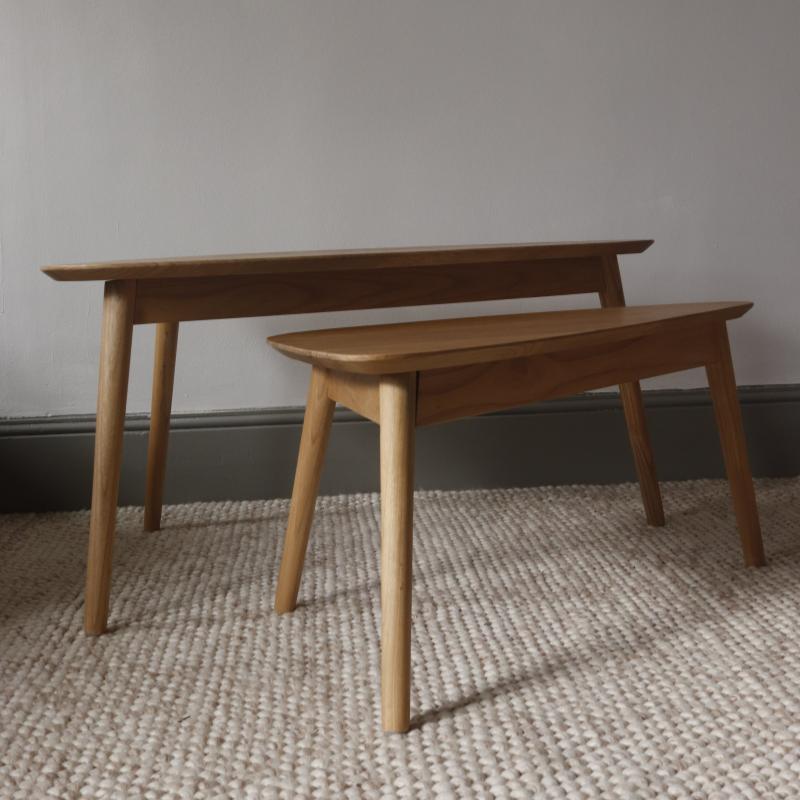 A light natural wood nesting table and bench set placed on a textured beige rug against a plain gray wall.