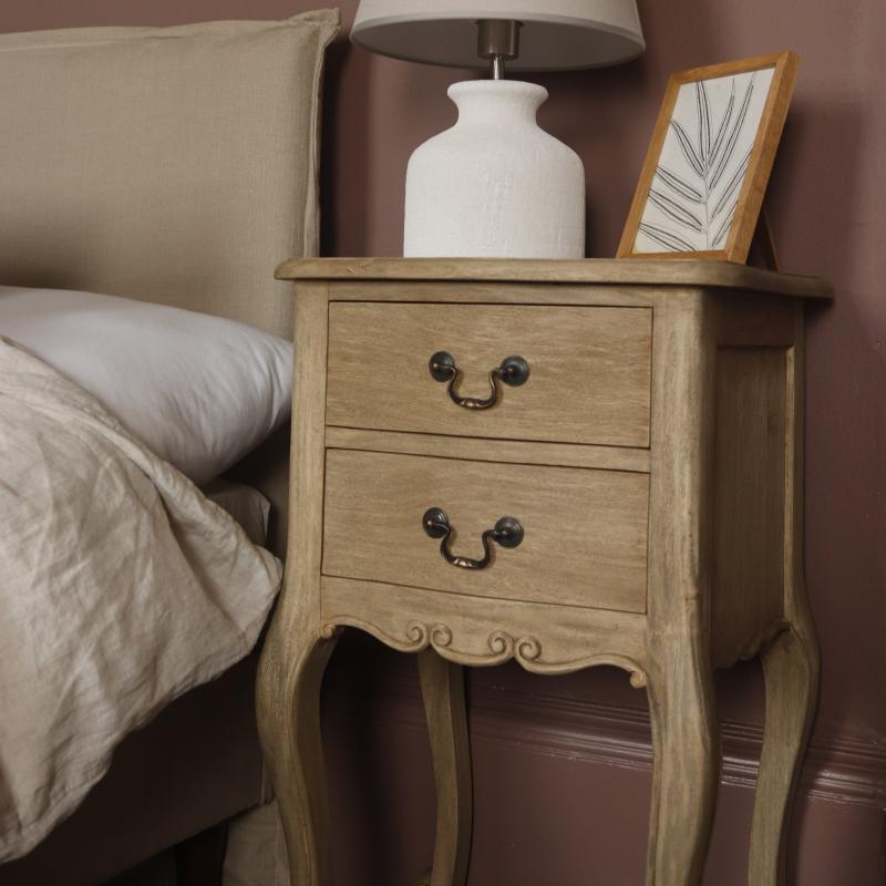 A light natural wood two-drawer nightstand with dark metal drawer pulls, styled next to a bed with beige bedding and a white table lamp on top.