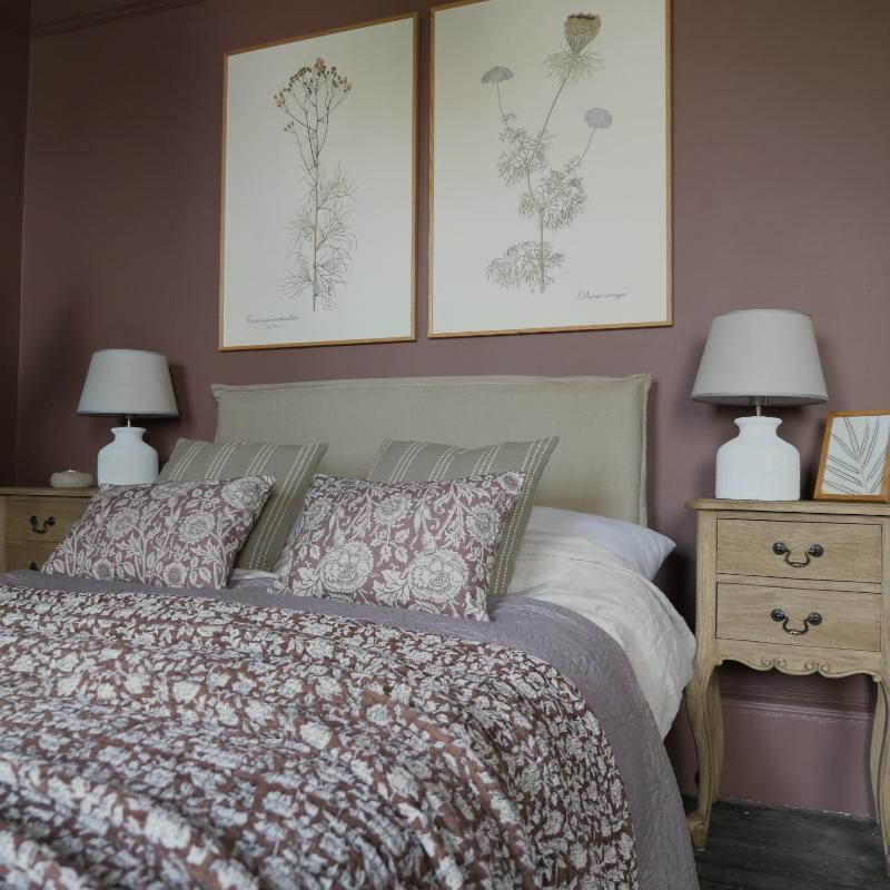 A styled bedroom scene featuring a bed with a floral patterned quilt and matching pillows, flanked by two light wood nightstands with white table lamps, set against a dusty rose wall with two botanical art prints above the headboard.