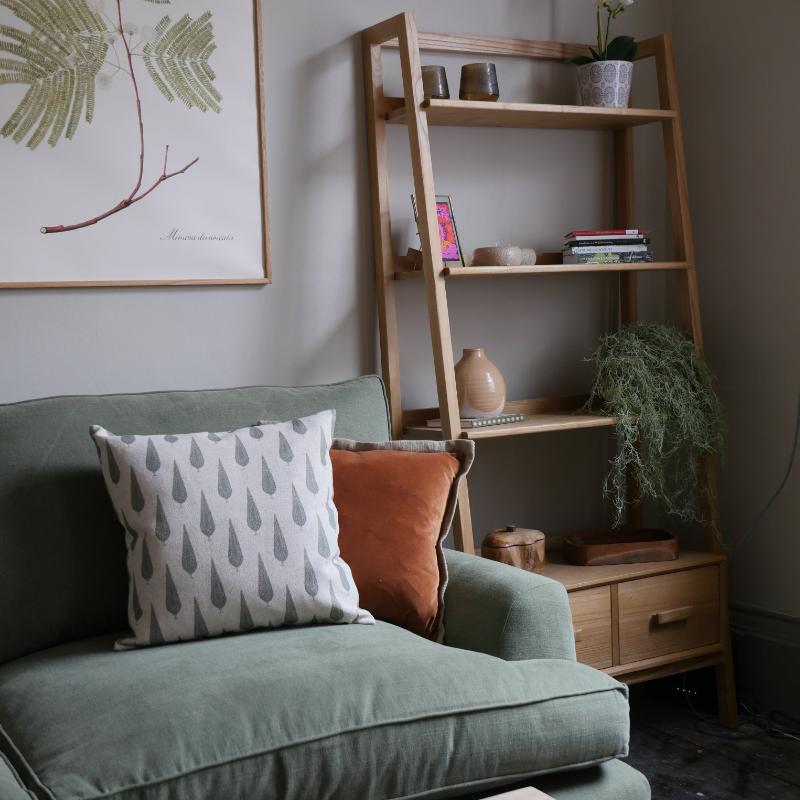 A styled interior corner showing a green upholstered armchair with patterned throw pillows, next to a tall wooden ladder shelf holding books, vases, and a trailing plant.