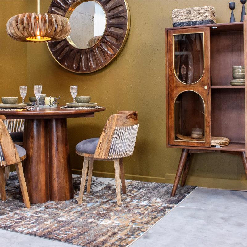 A styled dining room corner featuring a round wooden dining table, two woven-back chairs with gray cushions, a large sunburst wall mirror, a hanging wood pendant light, and a wooden display cabinet against an olive green wall.
