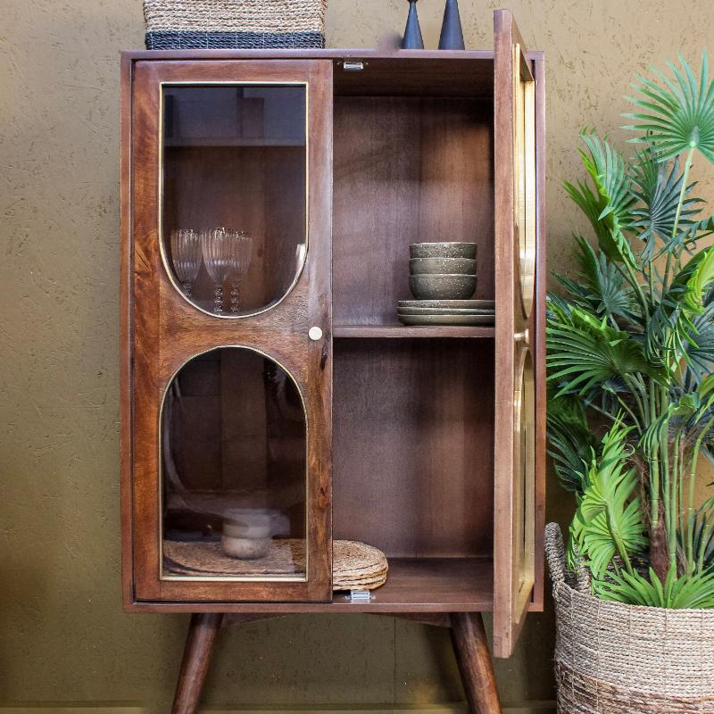 Mid-century modern wooden cabinet with glass-front upper door and open lower cabinet, shown in a styled interior setting with a potted plant and textured wall.