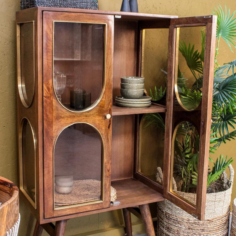 Mid-century modern wooden cabinet with glass doors and brass accents, styled in a home decor setting with plants and woven baskets.