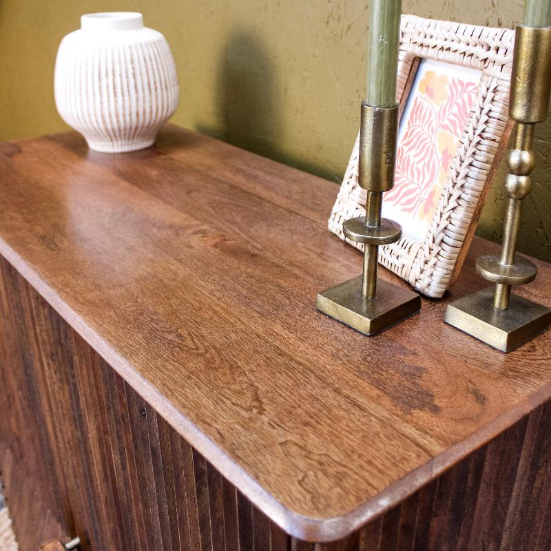 A close-up of a dark brown wooden sideboard or cabinet with a warm wood grain finish, styled with a white ribbed ceramic vase, a woven picture frame, and two brass candlesticks against a textured olive-green wall.