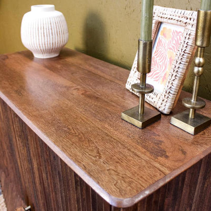 A close-up of a dark brown wooden sideboard or cabinet with a warm wood grain finish, styled with a white ribbed ceramic vase, a woven picture frame, and two brass candlesticks against a textured olive-green wall.