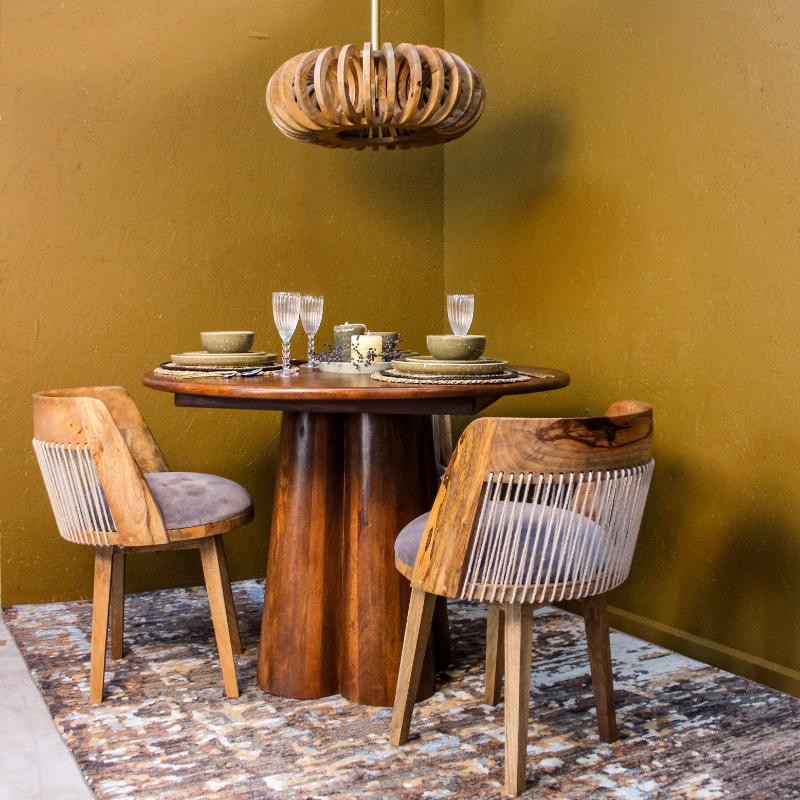 A rustic dining set featuring a round wooden table with a thick pedestal base, two matching wooden chairs with woven rope backs and gray cushions, and a matching wooden pendant light, set against a textured mustard yellow wall.