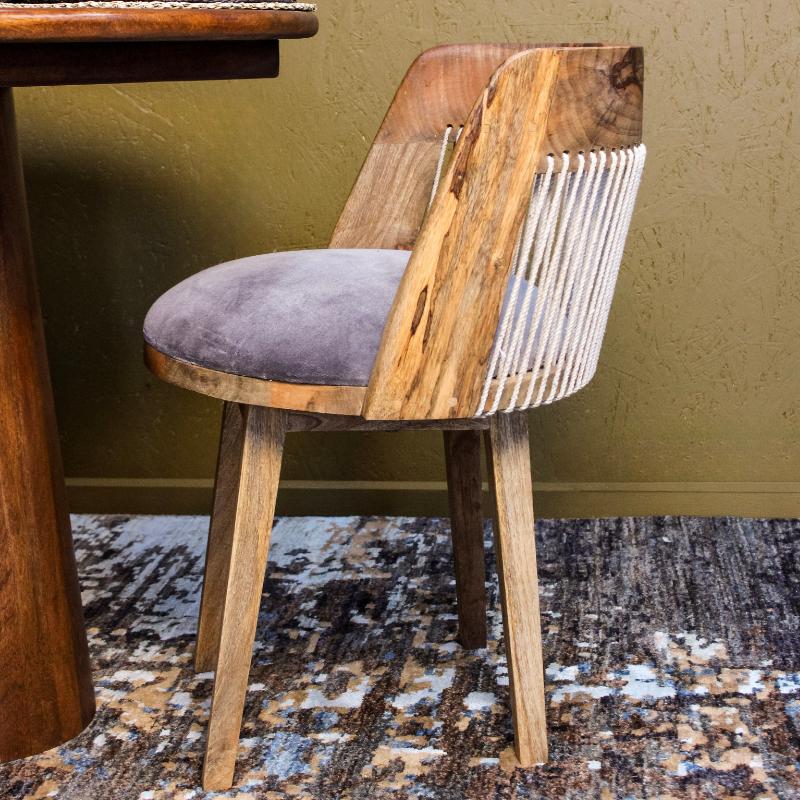 A wooden dining chair with a curved back, natural wood legs, and a gray velvet cushion, positioned next to a wooden table against a textured olive wall.