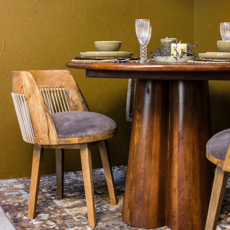 A rustic wooden dining chair with a gray velvet cushion sits beside a matching wooden table set with plates, a glass, and candles against a textured olive-green wall.