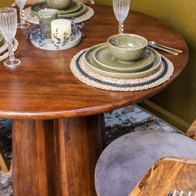 A styled dining table setting featuring a round wooden table with a matching green ceramic bowl and stacked plates on a woven placemat, alongside wine glasses, a candle, and part of a chair in a warm indoor setting.
