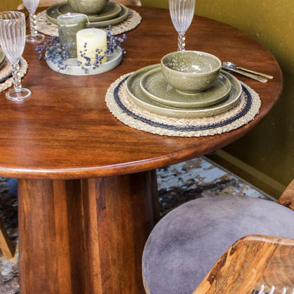 A styled dining table setting featuring a round wooden table with a matching green ceramic bowl and stacked plates on a woven placemat, alongside wine glasses, a candle, and part of a chair in a warm indoor setting.