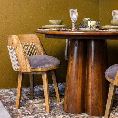 A rustic wooden dining table with a round top and thick pedestal legs, paired with two wooden chairs featuring woven rope backrests and gray velvet cushions, set against a textured olive green wall.