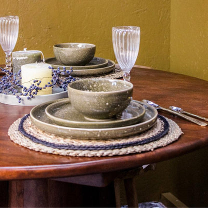 A styled dining table setting featuring speckled ceramic bowls and plates in a muted olive green and beige color, arranged on woven placemats with a candle, glassware, and silverware on a dark wood table against a mustard yellow wall.